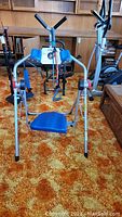 Front view of AB Flyer exercise machine showing metal frame, blue seat, arm handles, and digital counter.