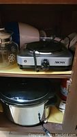 Wood cabinet shelf holding Aroma hot plate, slow cooker in lower section, with electric cords present.