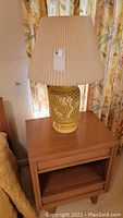 Pair of yellow ceramic table lamps with intricate cutout bases and pleated beige lampshades, shown on a wooden side table in front of floral curtains.