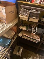 Photo showing a wooden shelving unit containing an inflator device, assorted cables, plastic storage containers, a metal lockbox on the floor, and various office supplies including envelopes and folders.