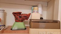 Green and red decorative item on stand next to plastic containers and cups on top shelf