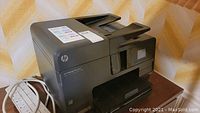 HP Officejet Pro 8610 printer showing front and paper tray on side of wooden surface with white power strip beside it