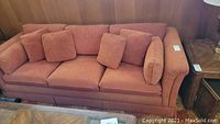 Full view of the rust-orange fabric sofa with three seat cushions and six matching pillows arranged on the backrest. The sofa has rolled arms and a skirted base.