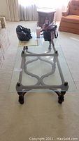 Full view of the coffee table in a living room with a glass top and ornate metal base. Bags and cup on top not included.