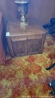 Photo of one wood side table showing patterned top with lamp and front storage doors on floral carpet