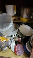 Various plastic storage containers stacked inside a cabinet including bowls and colanders, white and transparent containers with lids, along with metal pots and yellow handle items.