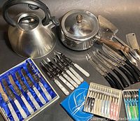 Photo showing the COPCO kettle, COPCO pot with wooden handle, various knives with black handles including serrated steak knives, mother of pearl-handled tea knives in original box, mid-century cocktail forks and espresso spoons in boxes.