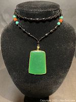 Front view of the necklace on a black stand showing the central green jade pendant with gold-tone framing and black oval beads with turquoise and brown bead accents.