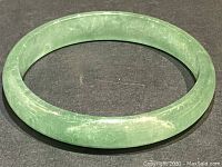 Solid circular bangle made from green jade against a dark surface, showing the natural color and texture of the jade.