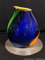 Full view of blue, green, and orange handformed glass vessel on round textured mat.