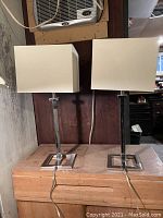 Pair of modernist metal base table lamps with cream square shades placed on wooden surface against dark wood background, showing full lamp height and square base detail.