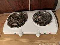 White double electric hot plate with two spiral heating coils, chrome burner rings, two white control knobs, and indicator lights on the front. Positioned on cardboard surface.