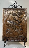 Front view of the wooden bas-relief carving plaque displayed on wrought iron stand. Shows detailed raven figure and carved textured background.