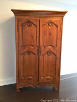 Full view of vintage wooden armoire showing front two doors with carved ornamental panels and decorative base.