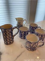 Porcelain pitchers and mugs with blue splatter design on white porcelain placed on marble surface.