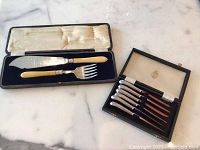 Overall view of two vintage flatware sets in their presentation cases: one fish serving set with ivory handles and one set of six silver fish knives with wooden handles.