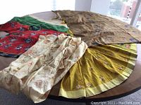 View of multiple swaths of silk fabric and one silk garment laid out on a round wooden table near a window with cityscape background.