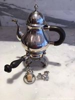 Overall view of the silver teapot on its stand with black handle and curved spout.