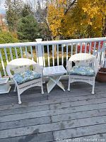 Two white wicker chairs with blue floral cushions placed on a wooden deck with white railings, accompanied by a white folding table between them.