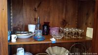 Shelf displaying crystal goblets, glass bottles, porcelain plates and bowls, hand blown glass bowl, and multiple champagne glasses.