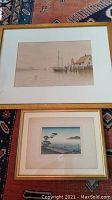 Two framed prints laid on patterned rug. Larger depicts a dock scene with sailboat and house, smaller shows a coastal landscape with trees and distant islands.