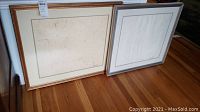 Two framed artworks showing nude figures in minimal line art style, placed on a hardwood floor. Both have wooden frames with matting and appear signed.