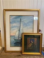 Two pieces of framed wall art together on a floor: larger watercolor of a sailboat signed Jane Collins, smaller framed painting on wood of a woman reading