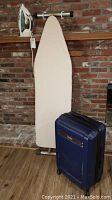 Full view of metal ironing board with beige cover, Sunbeam iron resting on the board, and blue Chaps rolling suitcase standing next to the board.