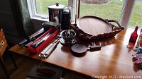 Wide view of kitchenware on wooden table including Revere Ware pot, loaf pans, graters, bottle opener, rattan tray, and cheese sets.