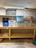 Photo showing six plastic storage bins on a wooden shelf, including translucent bins with blue and beige lids, plus black and blue plastic milk crates stacked to the side.