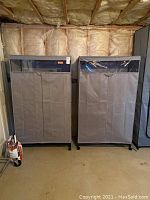 Two small rolling garment bags shown side by side in basement setting, both grey fabric with clear plastic windows and zipper openings on metal frames with wheels.