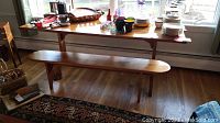 Photo shows handmade wooden table with various items on top and a long wooden bench beneath the table. The table has a natural wood finish with rounded edges and a central pedestal base.