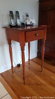 Side view of wooden side table showing drawer and turned legs with visible wear and stain on floor beneath.