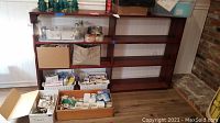 Wooden bookcase filled with various household items on shelves and boxes on floor in front; photograph displays scratches and general wear.