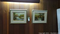 Pair of framed oil paintings hung on wood panel wall, both depicting landscape scenes with trees and water, signed A Lindsay