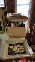 Stack of vintage wood frames inside a cardboard box with loose framed art featuring botanical motifs on table near door.