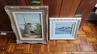 Two oil paintings on board side by side: one of a windmill in a natural landscape, the other a coastal scene with a white building. Both framed ornately.