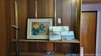 Three oil paintings on shelf, showing a framed abstract/surreal piece and two unframed landscape/seascape paintings on board.