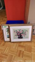 Two framed artworks leaning against a box, showing a floral still life and a tropical scene.