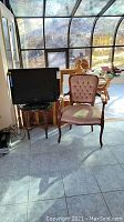 Room setting with Queen Anne style upholstered chair and vintage end table supporting a TV, natural light from window