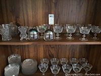 Full view of crystal sherry glasses, dessert plates and bowls, and cream and sugar set on wooden shelving.