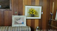 Two paintings hung on wood panel wall above plaid fabric furniture. One painting shows yellow flowers in vase, the other a still life with seashells and balls.