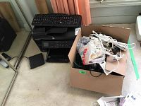 HP all-in-one printer with keyboard placed on top, next to a box containing various cables and accessories, all situated on a beige carpet.