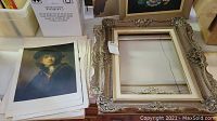 Top view photo showing stack of art prints lying next to two large ornate frames with intricate floral and scroll decorations.