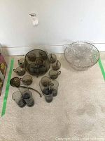 Two vintage glass punch bowl sets and matching glasses on floor. One set is clear patterned glass with ladle and glasses, the other smoky dark glass with gold rim glasses.