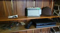 Wood shelf with vintage green Nordmende Royal radio, Vasotec remote controller, RCA VHS player, connecting cables, and a brown case on two shelves.