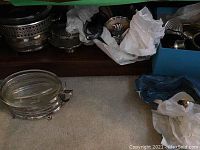 Wide shot of several silver plated serving bowls and items positioned on a carpeted floor with some parts wrapped in paper.