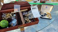 Full overview of brown velvet-lined jewelry box containing assorted vintage metal cufflinks, tie clips, and pins alongside a white open box with matching cufflink set.