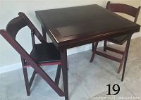 Front angled view of the red wood square folding table with two matching folding chairs with black leather seats.