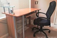 Photo shows a rectangular wood-finished office desk with silver metal accents on sides and vertical coverings, featuring shelves and a drawer. A black cushioned swivel office chair with armrests and wheels sits beside the desk. The desk is positioned near a window with horizontal blinds.